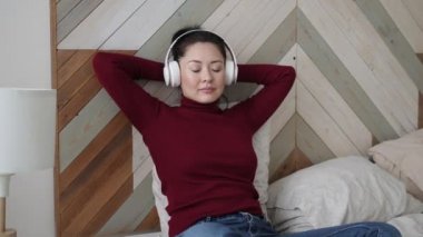 Genç Asyalı etnik kadın rahat bir yatakta, kulaklık takarak gözleri kapalı dinleniyor. Oldukça karışık ırk hanımı, rahat müzik dinlemeyi sever. Sesli meditasyon yapar. Evde stres hissetmez..