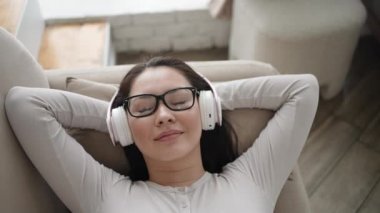 Kablosuz kulaklık takmış, rahat koltukta dinlenen çok ırklı genç bir kadın, Asyalı kız evde rahat bir müzik ve müzik dinlemeyi seviyor. Üst görünüm yavaş çekimde
