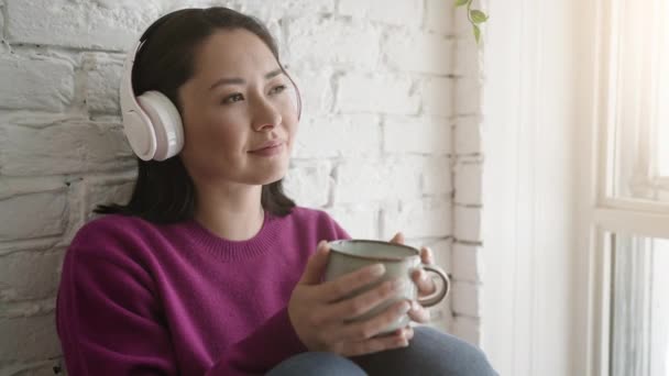 Atractiva joven mujer étnica sosteniendo taza de té o café bebiendo bebida caliente en casa. Joven asiática chica en auriculares escuchando música chill con bebida de la mañana mirando a través de la ventana y soñando — Vídeo de stock