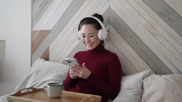 Ung asiatisk etnisk kvinde, der bruger smart phone surfing sociale medier, tjekker nyheder, spiller mobilspil eller sms 'er sidder på sengen derhjemme. Blandet race pige tilbringe tid med gadget teknologi. – Stock-video
