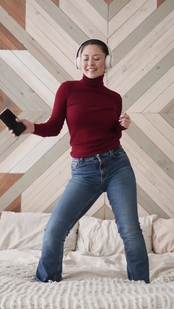 Überglückliche attraktive asiatische Frau, die zu Lieblingsdisco-Musik tanzt, auf dem Bett springt, die freie Wochenendfreizeit allein im Schlafzimmer genießt, fröhlich ist, während sie zu Hause das Freiheitskonzept feiert — Stockvideo