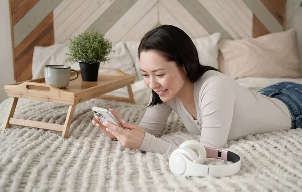 Wanita asia muda menggunakan telepon pintar berselancar di media sosial, memeriksa berita, bermain game mobile atau mengirim pesan duduk di sofa. Wanita ras campuran menghabiskan waktu di rumah dengan teknologi gadget sel. Stok Gambar