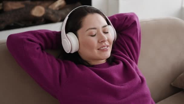 Joven mujer étnica escalofriante en cómodo sofá con los ojos cerrados con auriculares inalámbricos, mujer asiática disfruta escuchando música chill y sonido de audio relajante y meditando en casa. — Vídeo de stock