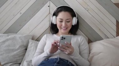 Genç Asyalı etnik kadın akıllı telefon kullanarak sosyal medyada sörf yapıyor, haberleri kontrol ediyor, mobil oyunlar oynuyor ya da koltukta oturmuş mesaj yazıyor. Sakin kız evde alet teknolojisiyle vakit geçiriyor..
