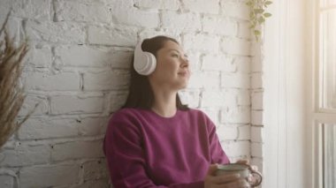 Evde çay ya da kahve içen çekici genç bir kadın. Kulaklıklı genç Asyalı kız sabah içkisiyle soğuk müzik dinliyor pencereden bakıp eğleniyor ve rüya görüyor.