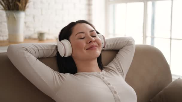 Ung asiatisk kvinna avkopplande på bekväm soffa med slutna ögon bär hörlurar. Ganska blandad ras dam gillar att lyssna chill musik ljud meditera känsla ingen stress hemma. — Stockvideo