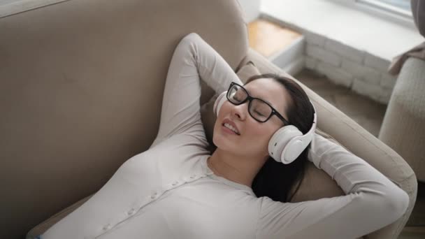 Joven mujer étnica escalofriante en cómodo sofá con los ojos cerrados con auriculares inalámbricos, mixta chica asiática disfruta escuchando música chill y sonido de audio relajante y meditando en casa. — Vídeo de stock