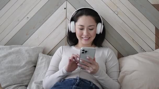 Mujer étnica asiática joven utilizando el teléfono inteligente navegar por las redes sociales, comprobar las noticias, jugar juegos móviles o mensajes de texto sentado en el sofá. Chica tranquila pasar tiempo en casa con la tecnología gadget. — Vídeos de Stock