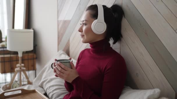 Joven mujer de ensueño étnica asiática en auriculares que sostienen la taza disfrutar de la bebida de té favorita bebida de café de la mañana mientras se sienta en casa en el dormitorio antes de empezar a trabajar. Ruptura, pausa, concepto de ensueño. — Vídeo de stock