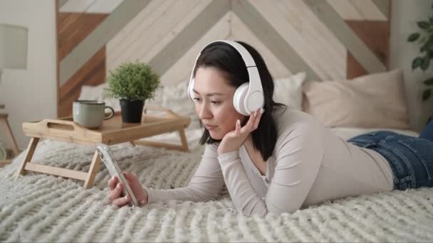 Młoda azjatycka etniczna kobieta korzystająca ze smartfona surfującego po mediach społecznościowych, sprawdzająca wiadomości, grająca w gry mobilne lub wysyłająca SMS-y na sofie. Spokój dziewczyna spędza czas w domu z technologii gadżet. — Wideo stockowe