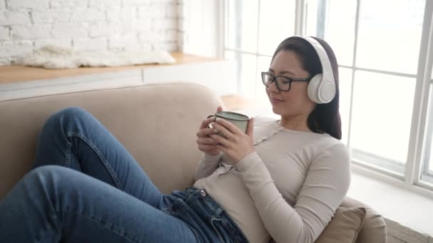 Молода етнічна жінка охолоджує на зручному дивані з закритими очима в бездротових навушниках, азіатська змішана расова дівчина насолоджується прослуховуванням холодної музики та звуку, що розслабляється вдома — стокове відео