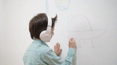 Kulaklıklarla resim çizen sevimli, komik okul çocuğu çocuk ressam tebeşirle boyama resmi, beyaz duvara boya kalemi, evde yaratıcı sanat aktivitelerinden hoşlanan zeki çocuk, çocuk gelişimi konsepti.