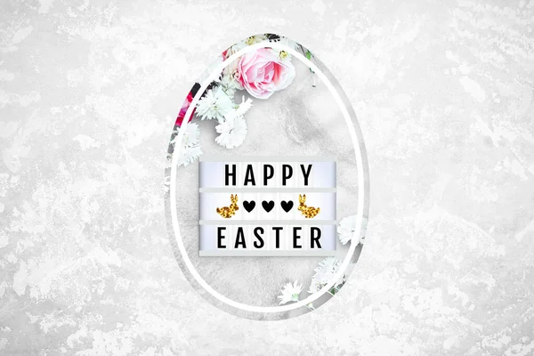 Lightbox and flowers in Easter egg shaped hole on a concrete background. Red and pink roses, white daisies. Lightbox with the lettering Happy Easter, hearts, bunnies. Top view, close up, copy space
