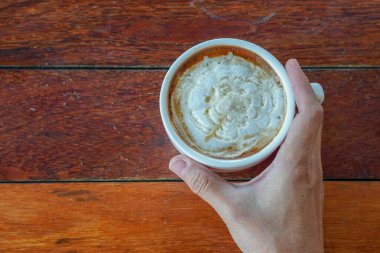 Üst manzara, sağ el sıcak karamelli macchiato kahveyi beyaz seramik bardakta ahşap bir masada tutuyor, yiyecek, içecek, kopyalama alanı.