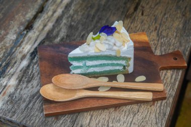cake with three layer of cake and three layer of white cream, on top by coconut and a butterflypea flower, sprinkle with almond slices which place on brown chopping board and two light brown wooden spoons, on wooden table background