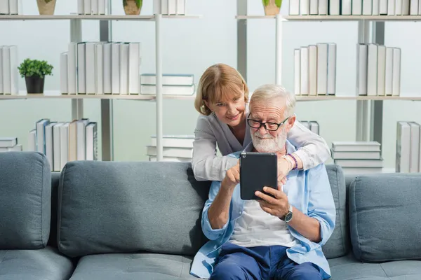 Yaşlı, üst sınıf aile çifti, günlük kıyafetlerini giyer ve akıllı tablet kullanır, gülümser, mutlu ve romantik bir şekilde oturma odasında gülümserler..
