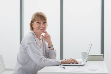 Gündelik kıyafet giymiş yaşlı bir kadının portresi. Oturma ve iş hayatı modern çalışma odasında gülümseme ve mutlulukla evde çalışıyor..