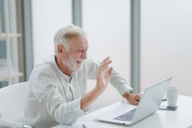Beyaz giysili kıdemli yaşlı adam, modern bir çalışma odasında, evde oturarak ve ekip olarak online video görüşmeleri yaparak çalışıyor..