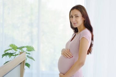 Hamile kadın portresi, gündelik kıyafetlerle modern yatak odasında çocuğunu karnında tutuyor ve ayakta tutuyor..