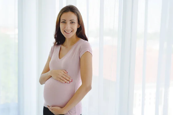 Hamile kadın portresi, gündelik kıyafetlerle modern yatak odasında çocuğunu karnında tutuyor ve ayakta tutuyor..