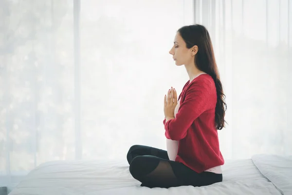 Hamile kadın portresi, gündelik kıyafetlerle yatakta oturup modern yatak odasında yoga yapıyor..