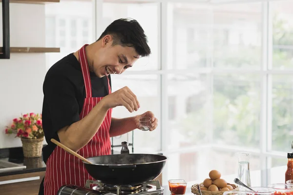 Modern mutfak odasında önlük giymiş Asyalı genç bir adam kahvaltı için yemek pişiriyor..