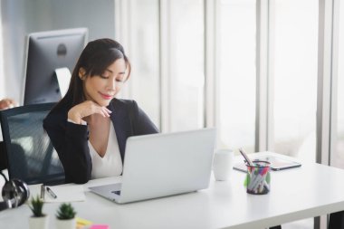 Resmi takım elbiseli Asyalı bir iş kadını modern ofis işyerinde gülümseyen ve bilgisayarını daktilo ederek çalışıyor..