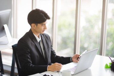 Business Asian man in formal manager looking suit working using smart phone with smiling and confident emotion in modern interior office workplace.