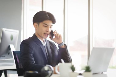 Business Asian man in formal manager looking suit working using smart phone with smiling and confident emotion in modern interior office workplace.
