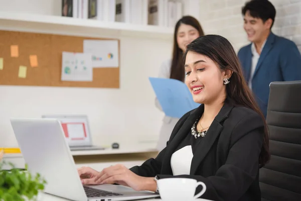 Young business Asian woman in formal outfit using laptop for working with confidence in modern workplace with colleagues in background.