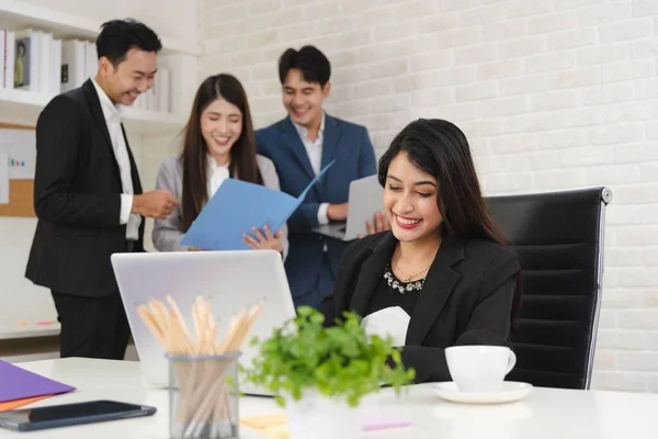 Young business Asian woman in formal outfit using laptop for working with confidence in modern workplace with colleagues in background.