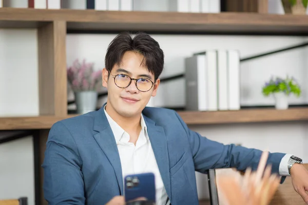 Portrait of young Asian business man in formal suit using smart phone in modern workplace.