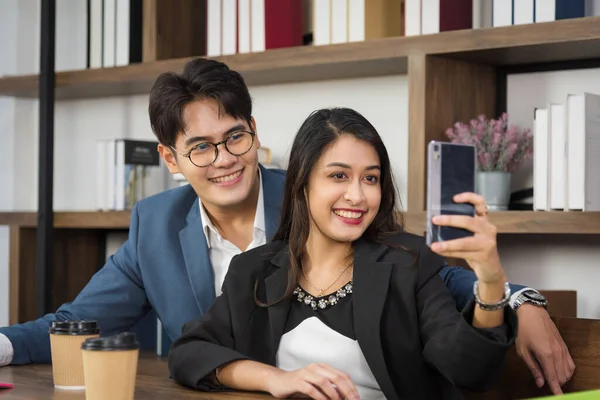 Portrait of young Asian business people in formal suit using smart phone for selfie in break time in modern workplace.