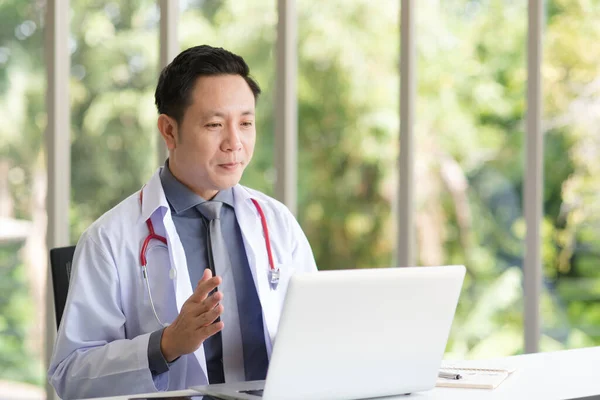 Asian Doctor man in white professional lab coat uniform sitting and using laptop in modern workplace office.