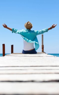 Çocuk denizin önünde şapkasıyla iskelede oturuyor.