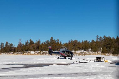 Helikopter kış günü helikopter pistinden havalanıyor. Etrafı ağaçlar ve karlarla çevrili.