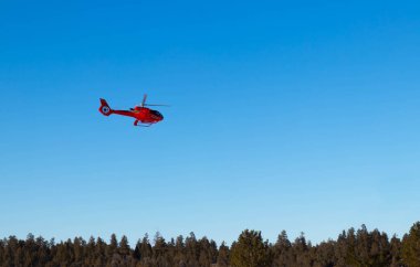 Açık mavi gökyüzünde, ormanda uçan kırmızı helikopter.