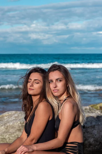 Two lesbian women hugging on the beach in bikinis between rocks and ocean — Stock Photo, Image