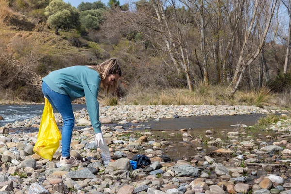 Woman crouching down to pick up a plastic bottle found among the river stones