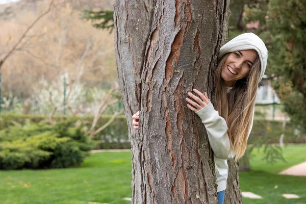 Leende och glad ung kvinna bakom trädstammen med en tröja huva — Stockfoto