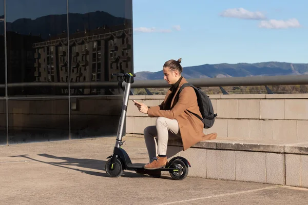 Moderner junger Mann mit Dutt-Frisur textet auf dem Smartphone gefesselt auf einer Bank mit nachhaltigem Elektroroller — Stockfoto