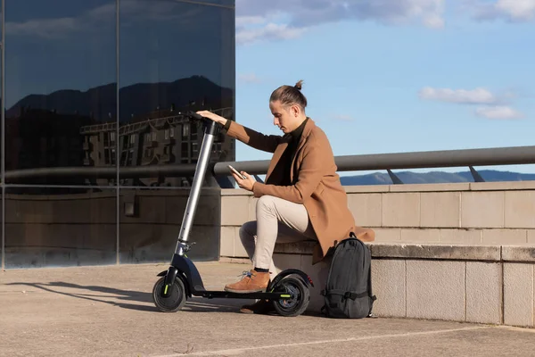 Genç modern adam bankta oturuyor ve elektrikli scooterıyla cep telefonu kullanıyor. Yeşil ve temiz enerji kullanıyor.