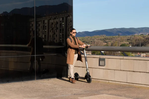 Jeune homme dans la vie de la ville avec un scooter électrique et tasse de café sur sa main marchant autour de la ville avec la montagne derrière — Photo