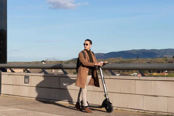 Arka planda güzel bir manzara ve elektrikli scooterıyla şehirde dolaşan genç bir işadamı. — Stok fotoğraf