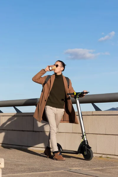 Junger Geschäftsmann trinkt Kaffee auf einer Tasse, während er mit seinem Elektroroller unter blauem Himmel durch die Stadt scrollt — Stockfoto