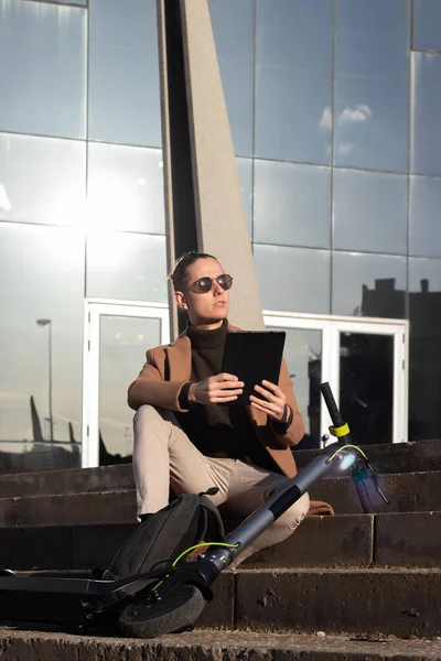 Vertikale Ansicht eines jungen, gut aussehenden Geschäftsmannes, der auf seinem Tablet im Treppenhaus mit einem Elektroroller ein E-Book liest — Stockfoto