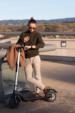 Elektrikli scooterla gidip gelen genç ve yakışıklı işadamının dikey fotoğrafı ve akıllı saat üzerinden konuşması.