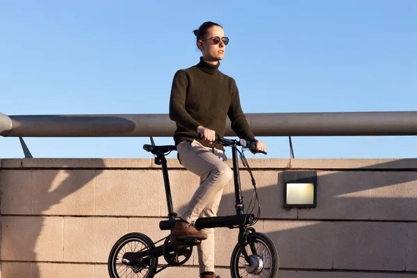 Jeune homme sur des lunettes de soleil chevauchant un vélo électrique autour du parc de la ville sous le ciel bleu avec la lumière du coucher du soleil sur son visage — Photo