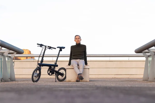 Jovem sentado em um banco de parque com sua bicicleta de bateria no lado olhando para a câmera sorrindo com espaço de cópia — Fotografia de Stock