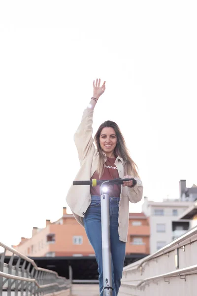 Vertikale Aufnahme einer jungen Frau, die Spaß mit ihrem Elektroroller durch die Stadt hat. Erhobener Arm als Glückszeichen — Stockfoto